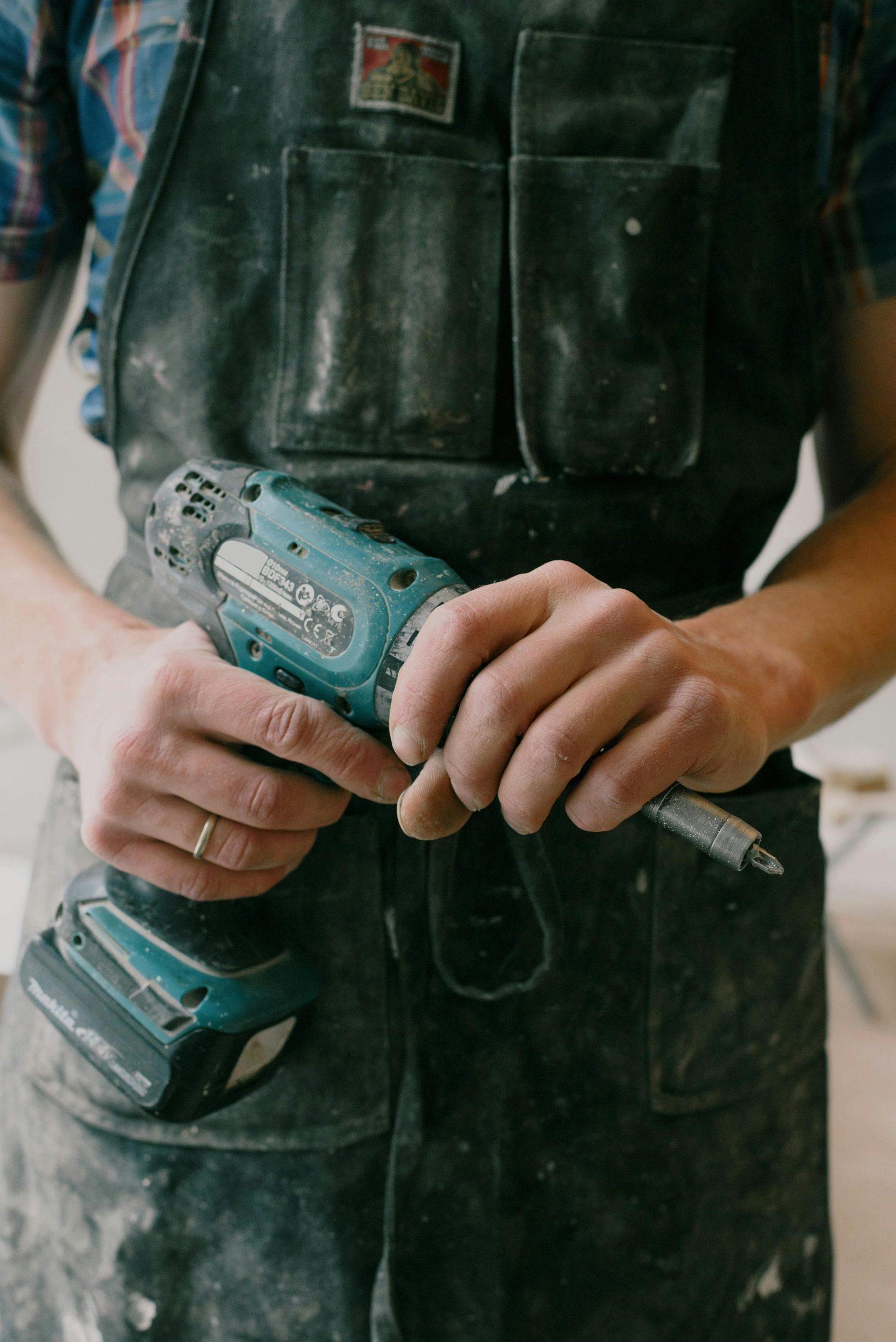 A man in coveralls seen from the shoulders down holding a power drill - Visit Phaselectric.com.